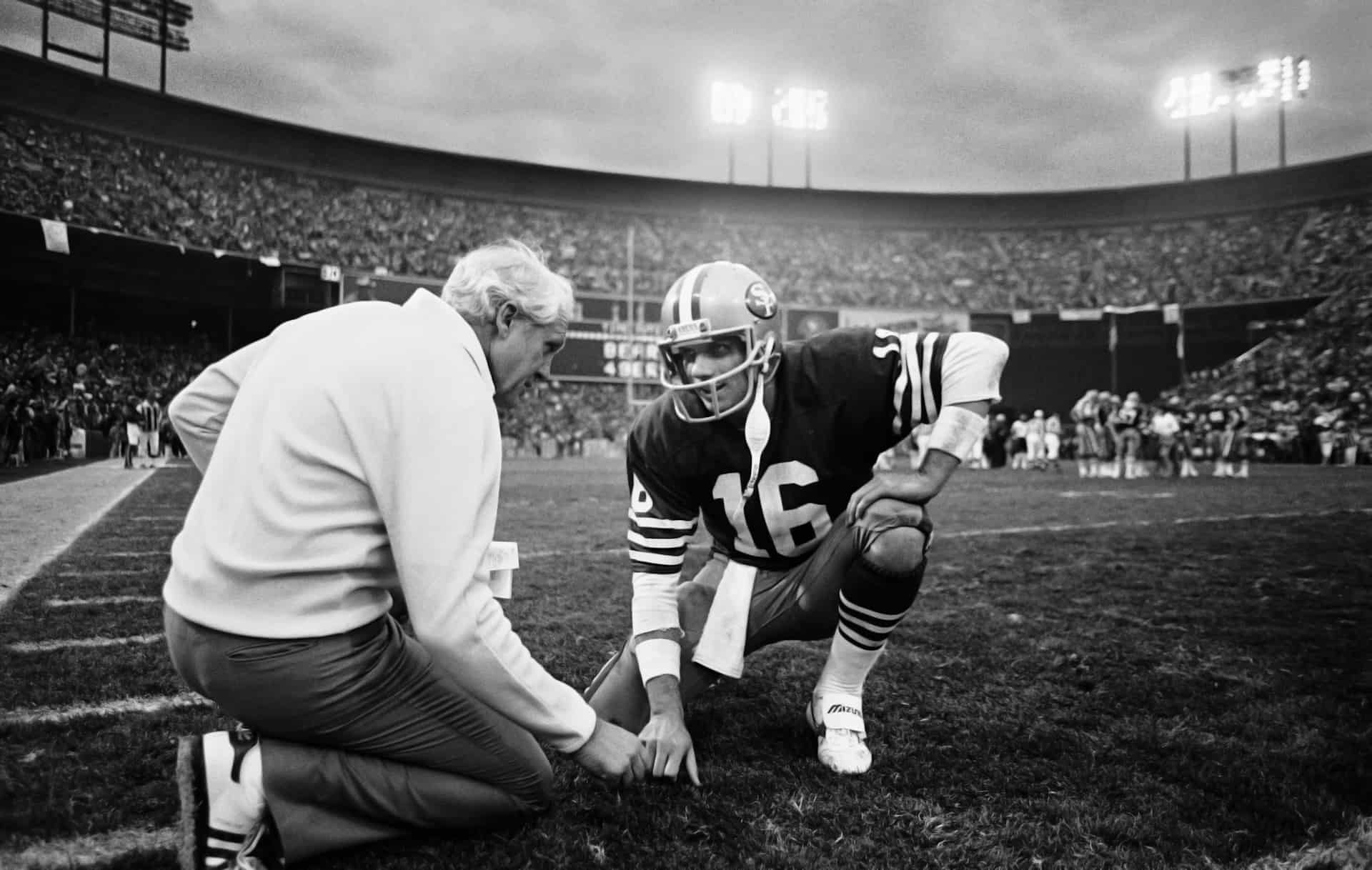 Joe Montana kneels at the field's edge to coordinate with the team's coach.